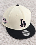 Gorra crema con visera plana negra, bordado LA negro con contorno rosa y parche lateral